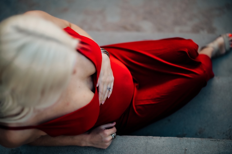 A woman is pregnant and places her left hand on her belly while she sits on a concrete step. She wears an elegant red dress.