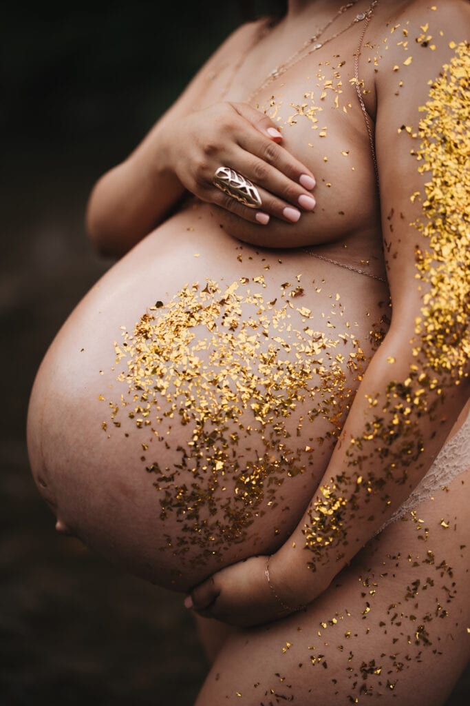 A woman holds her bare belly, she is pregnant, her body is covered in gold glitter